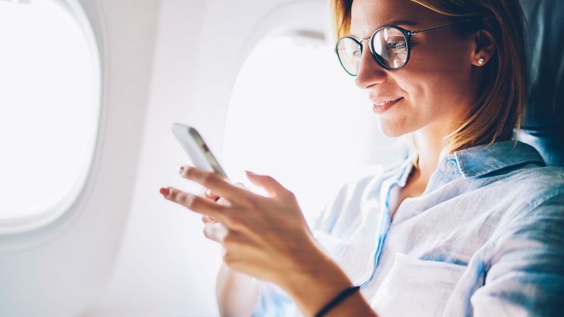 Eine Frau Sitzt am Fenster im Flugzeug und lächelt in ihr Smartphone.